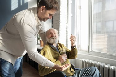 A young man giving medicine to an older man in a wheelchair. Caregiving featured image