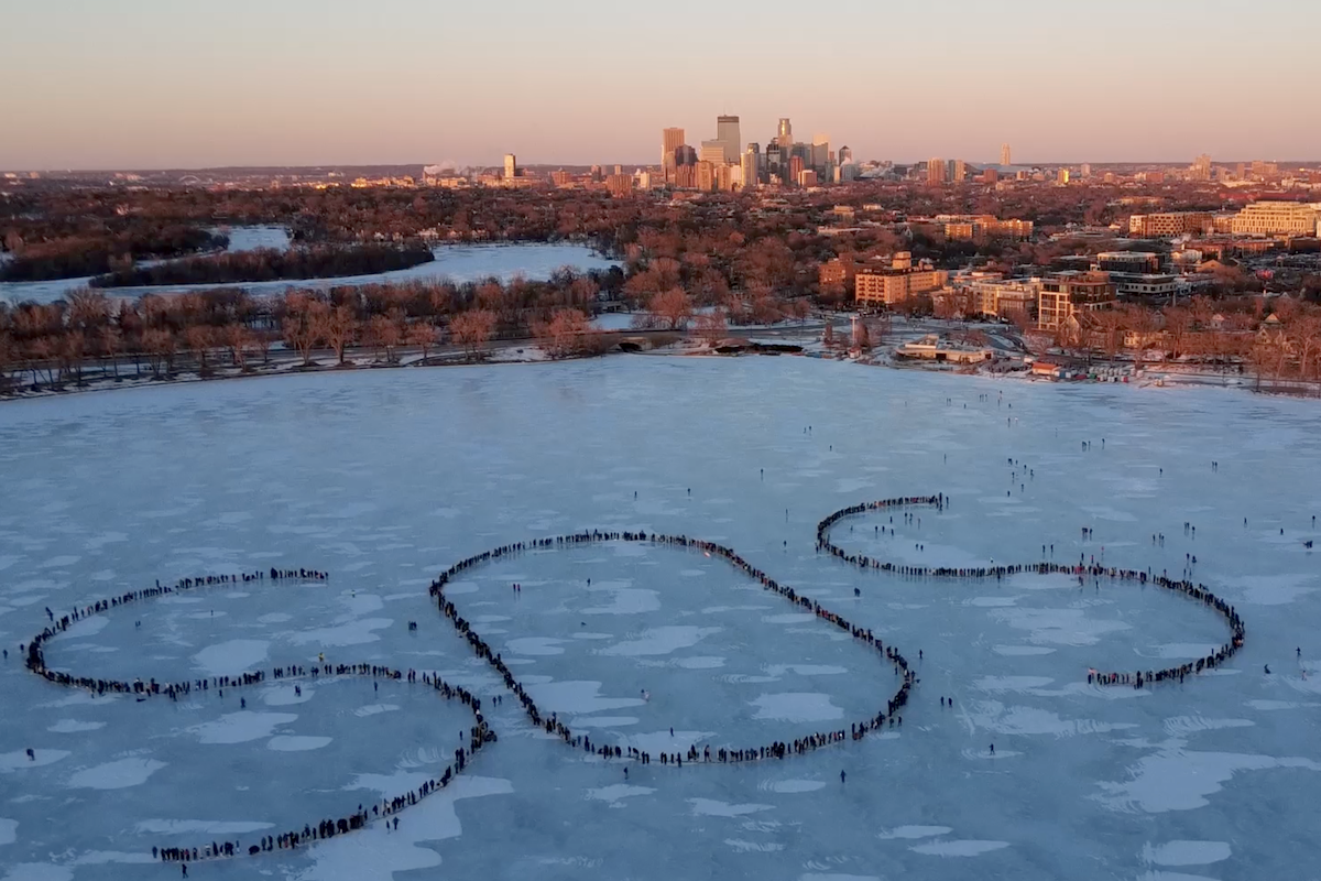 Hundreds of people form a human distress signal on Bde Maka Ska, a frozen lake in Minneapolis, with the city's skyline in the background.