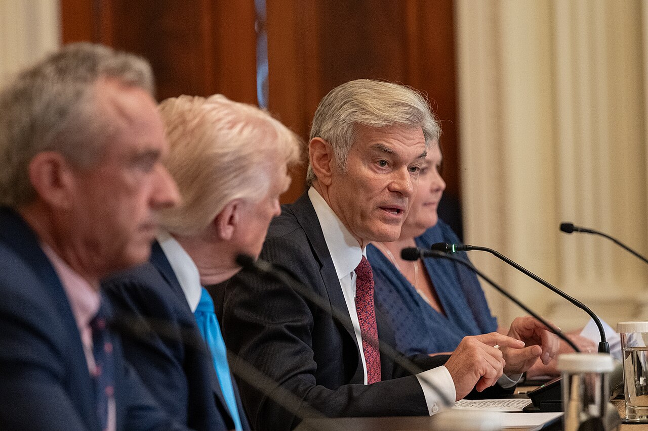 CMS Administrator Dr. Mehmet Oz talks into a microphone next to President Trump and HHS Secretary Robert F. Kennedy Jr. in July 2025