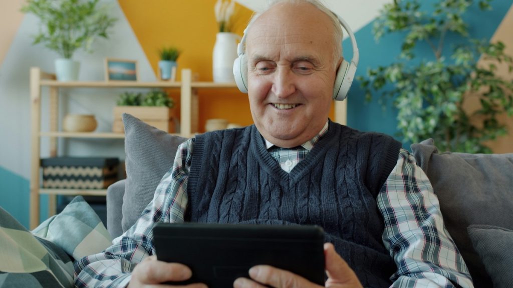 older adult smiling using a tablet and wearing headphones. AI and aging