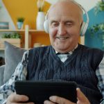 older adult smiling using a tablet and wearing headphones. AI and aging