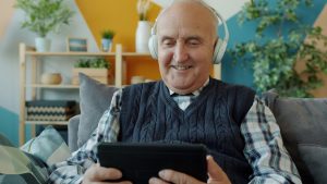older adult smiling using a tablet and wearing headphones. AI and aging