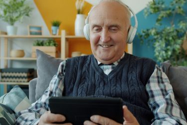 older adult smiling using a tablet and wearing headphones. AI and aging