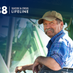 An older man with a glorious mustache driving a tractor with the 988 Suicide & Crisis Lifeline logo in the upper left corner.