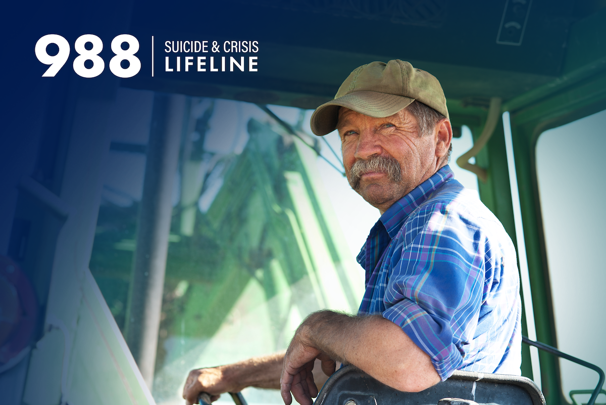 An older man with a glorious mustache driving a tractor with the 988 Suicide & Crisis Lifeline logo in the upper left corner.